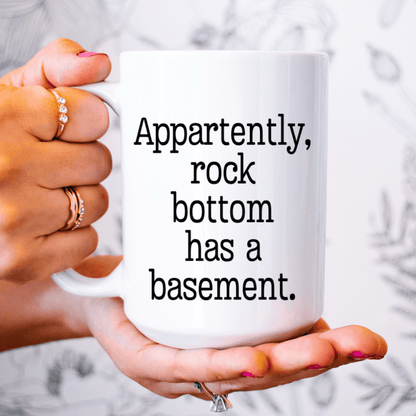 White coffee mug with Apparently Rock Bottom Has a Basement printed in black, perfect for sarcastic humor lovers.
