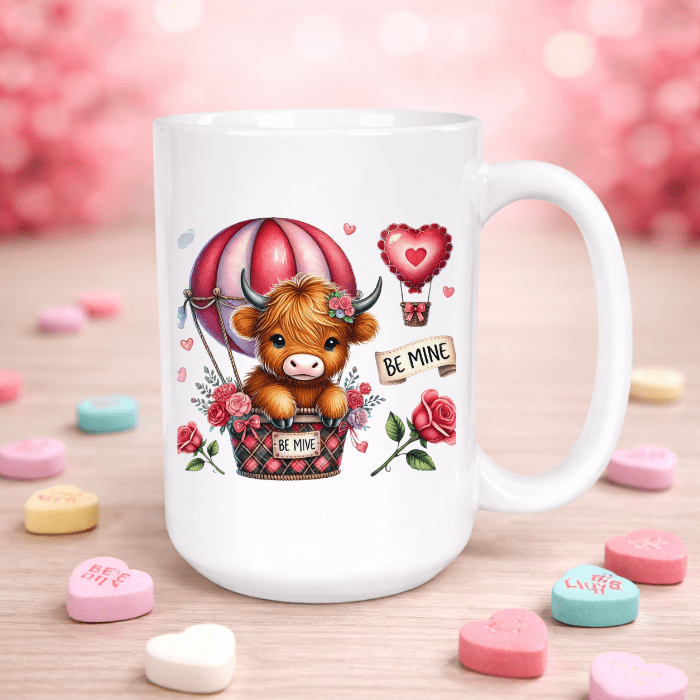 White mug with a cartoon cow in a hot air balloon surrounded by hearts and 'Be Mine' signs on a wooden surface with candy hearts.