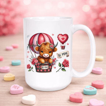 White mug with a cartoon cow in a hot air balloon surrounded by hearts and 'Be Mine' signs on a wooden surface with candy hearts.