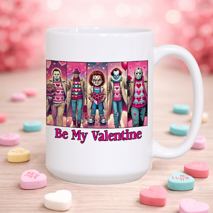 White mug with Valentine-themed design featuring characters in sweaters and 'Be My Valentine' text, surrounded by heart-shaped candy on a wooden surface.