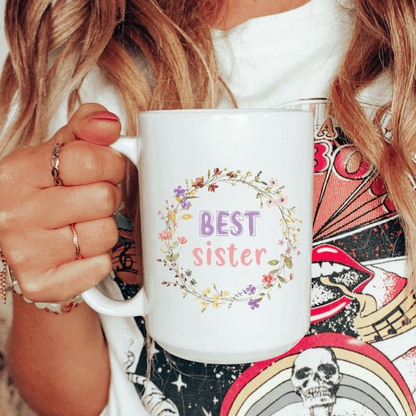 Person holding a white mug with 'BEST sister' text and floral design.