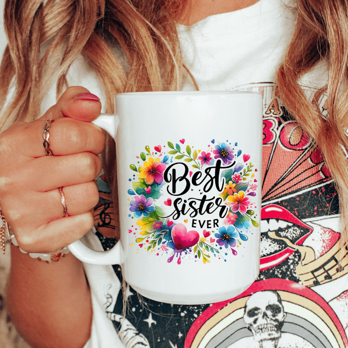 Person holding a white mug with colorful 'Best Sister Ever' design