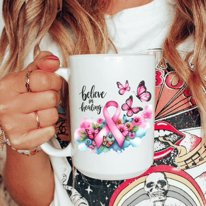 Person holding a mug with a pink ribbon and butterflies design, featuring the text 'believe in healing'.