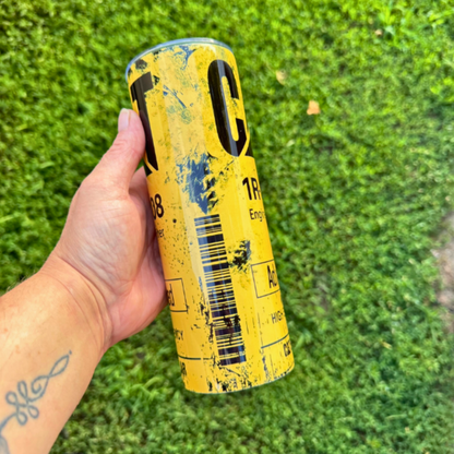 A hand holding a yellow and black cylindrical can with a distressed, weathered design, set against a background of green grass.