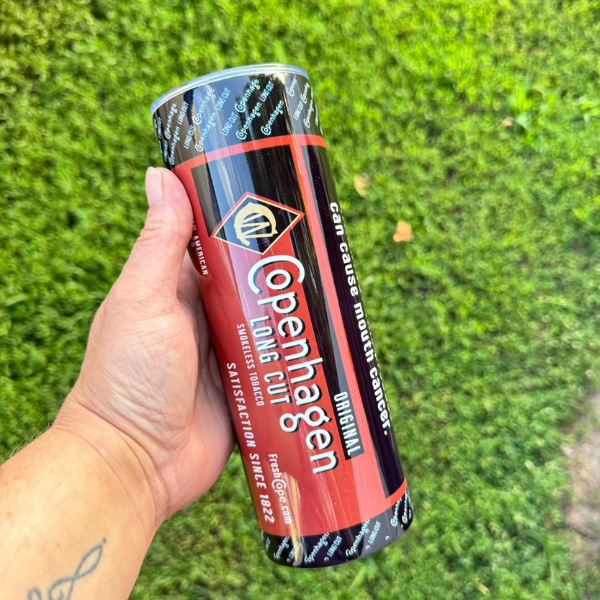 Hand holding a can of Copenhagen Original against a grass background