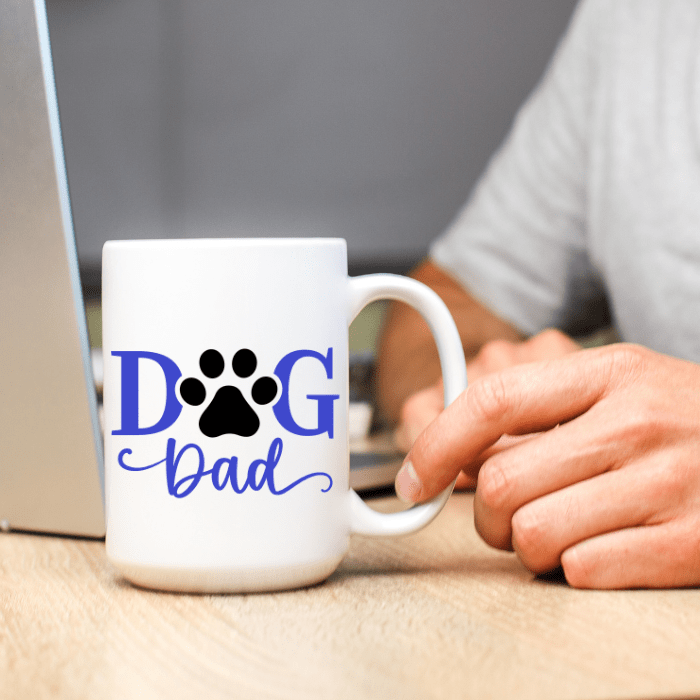 White mug with 'DOG Dad' text and paw print held by a person at a desk.