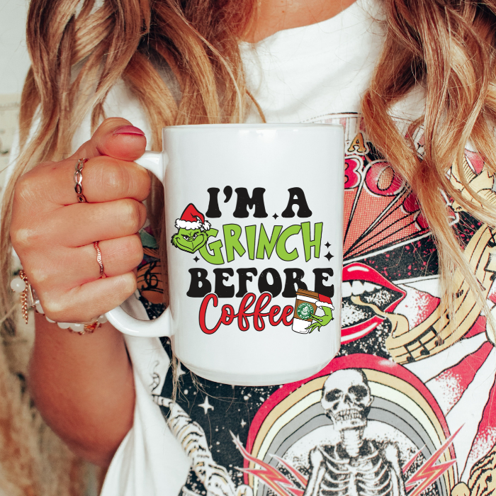 Person holding a white mug with 'I'm a Grinch before coffee' text, wearing a colorful patterned shirt.