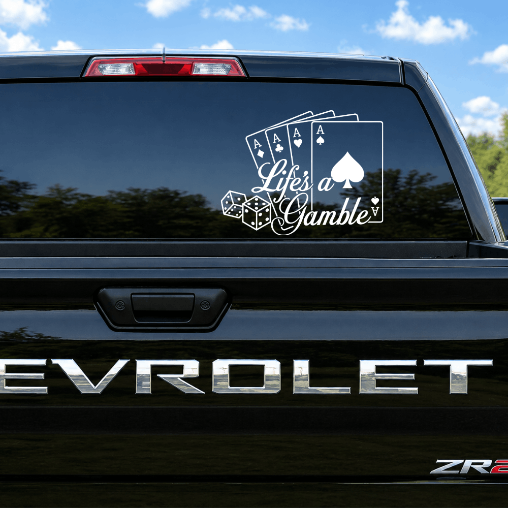 A white vinyl decal with the phrase 'Life is a Gamble' and playing card graphics applied to the back window of a black Chevrolet truck, shown outdoors under a blue sky.