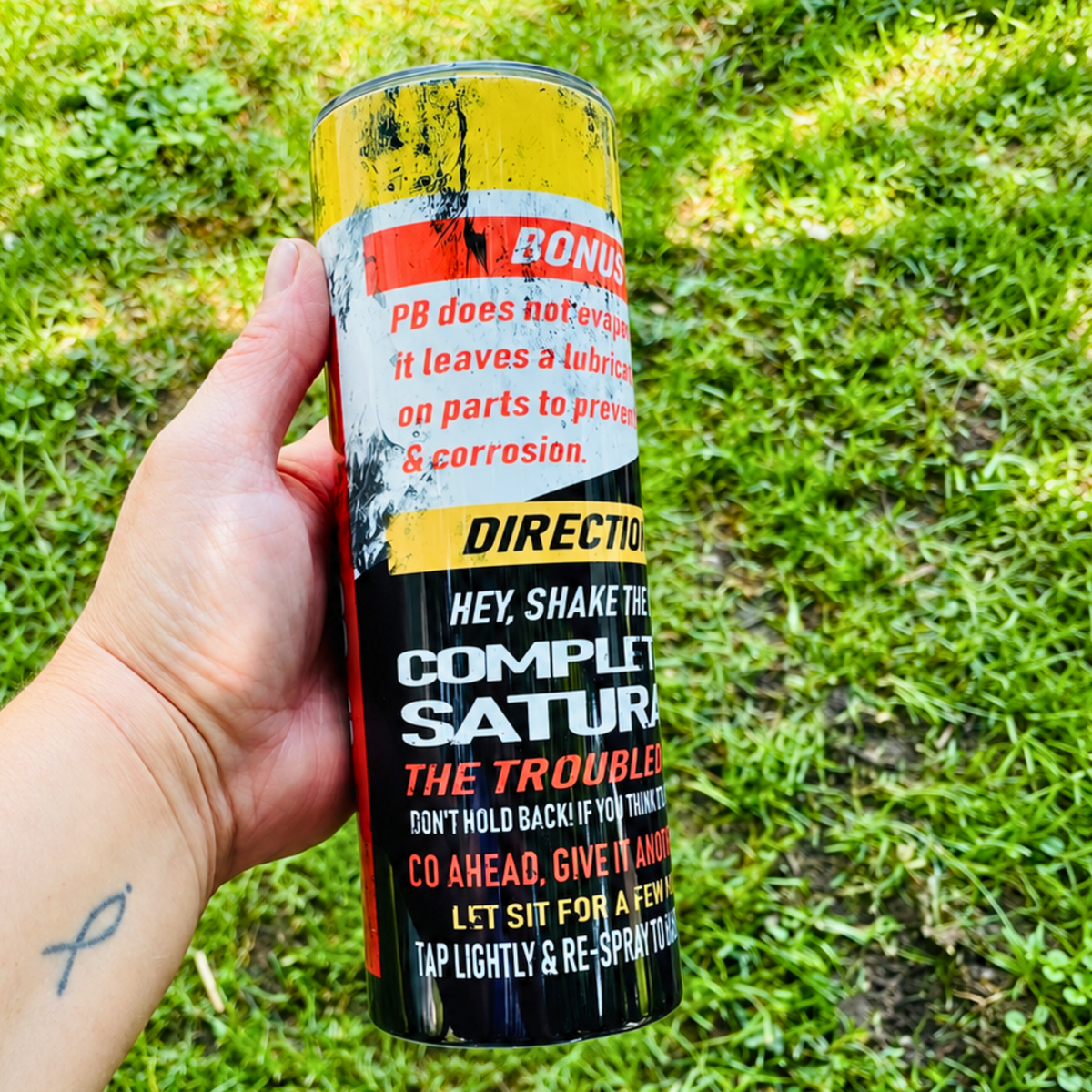 A hand holding a can of PB Complete Satur Lubricant outdoors on a grassy lawn. The can features a black, yellow, and red label with instructions and product details.