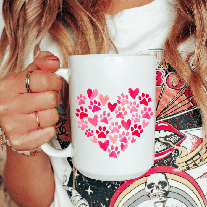 White mug with pink heart and paw print design held by a person with a colorful background.