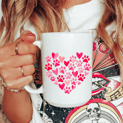 White mug with pink heart and paw print design held by a person with a colorful background.