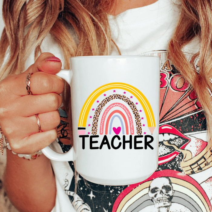 Person holding a white mug with a rainbow and heart design labeled 'TEACHER'.