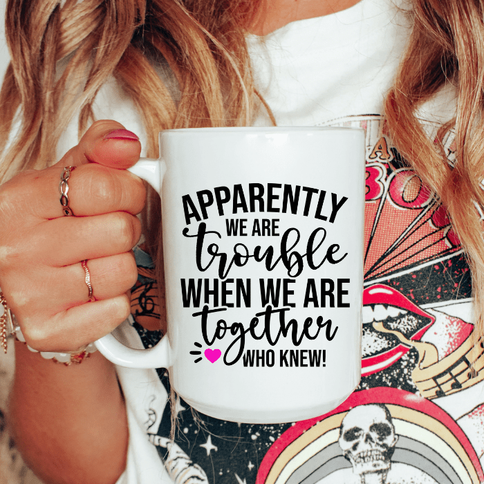 Person holding a mug with text 'Apparently we are trouble when we are together who knew!' against a colorful background.