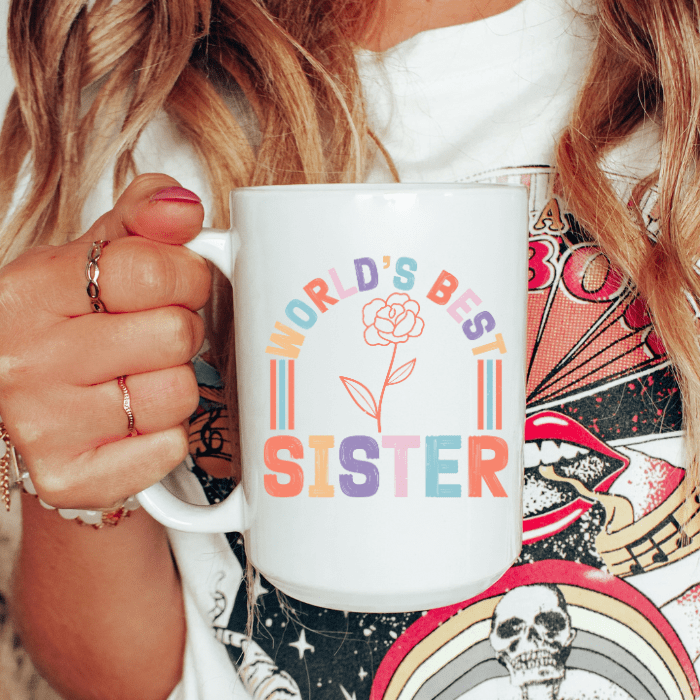 Person holding a white mug with 'World's Best Sister' text and flower design.